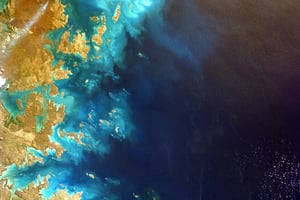 Satellite view of coastal landmasses meeting deep blue ocean waters, with swirls of turquoise and tan indicating shallow reefs and sediment patterns. The image highlights Earth’s natural beauty and environmental complexity.  Credit: Photo by NASA on Unsplash
