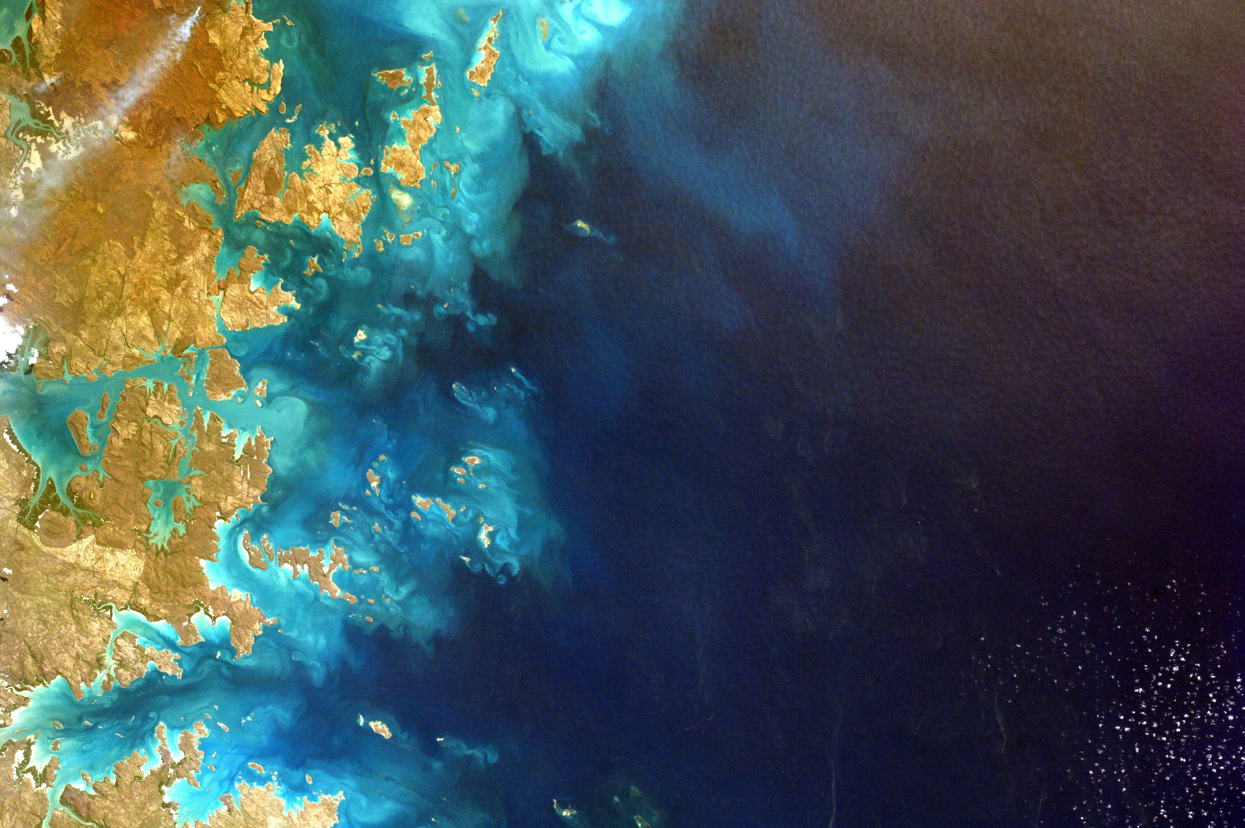 Satellite view of coastal landmasses meeting deep blue ocean waters, with swirls of turquoise and tan indicating shallow reefs and sediment patterns. The image highlights Earth’s natural beauty and environmental complexity.  Credit: Photo by NASA on Unsplash