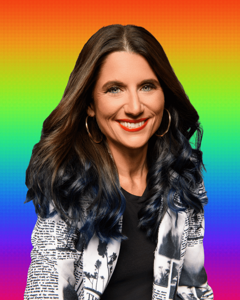 A confident woman with long, dark wavy hair featuring subtle blue highlights smiles warmly against a vibrant rainbow gradient background. She’s wearing large gold hoop earrings, a black shirt, and a statement blazer with black-and-white newspaper-style print and palm tree imagery. Her look is polished, creative, and bold—reflecting a dynamic personal brand.